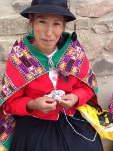 Peruvian woman in traditional dress knitting while sitting on a stone step