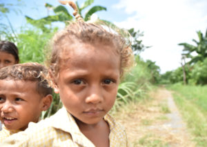 Young girl in Timor Leste on side of dirt road smiling at camera with palm trees in the background