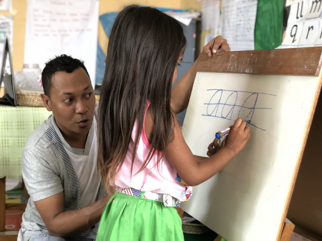 Young Timorese girl at school at a flip chart writing with a teacher crouched helping her