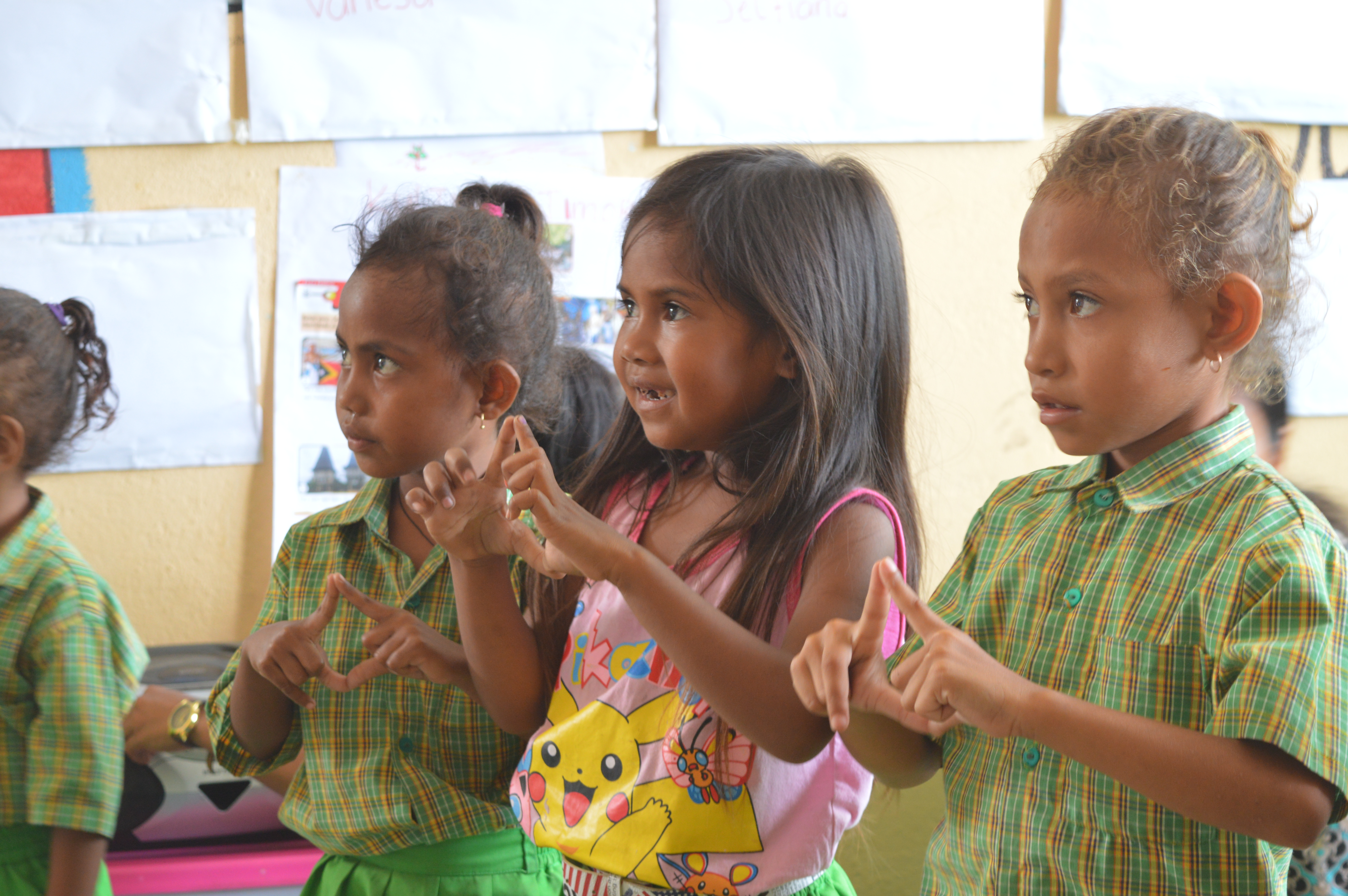 Three primary school children looking away from the camera are singing with their hands in shape of diamond in front of them.
